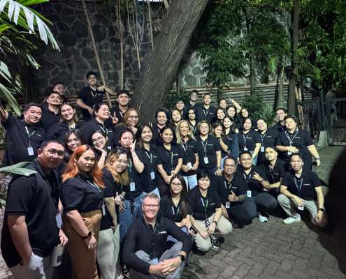 Group of about 40 coworkers posing for a team photo outdoors at night, all wearing black polo shirts and ID lanyards, with trees and a parked vehicle in the background.