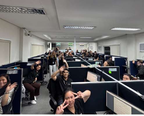 Group of call center agents in cubicles waving at the camera in a busy office environment with many desks.